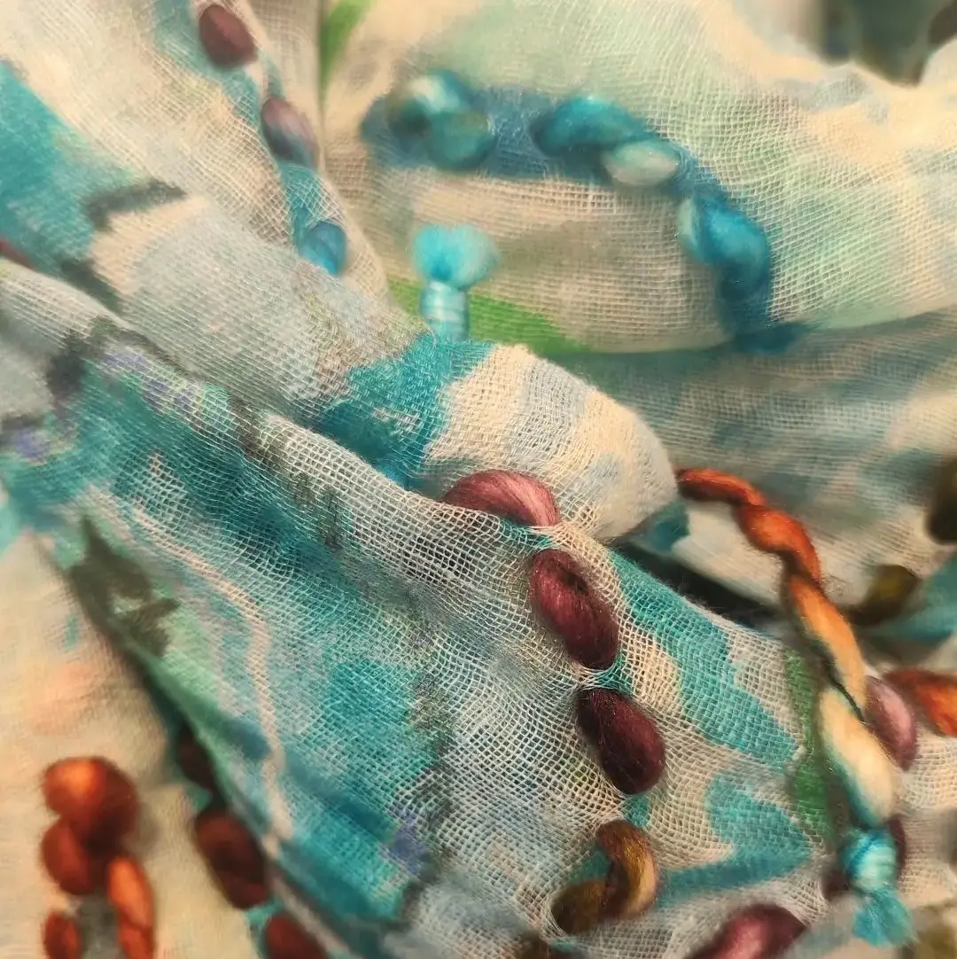 Primer plano de pañuelo artesanal de Cado Complementos en seda y modal turquesa con bordados en relieve de lana multicolor. Texturas textiles de alta gama.